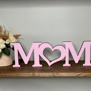May include: Pink wooden letters spelling "MOM" with a heart cutout between the "M"s. The letters are on a wooden shelf.