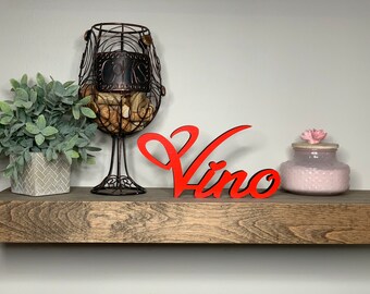 May include: A red wooden sign with the word "Vino" in a cursive font, displayed on a wooden shelf with a wine cork holder, a potted plant, and a pink decorative jar.