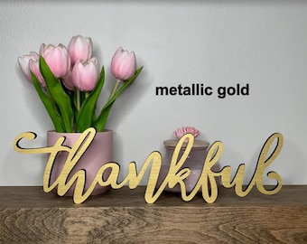 May include: A metallic gold script sign featuring the word thankful in a cursive font. The sign is positioned in front of a pink vase containing light pink tulips and a small purple jar with a wooden lid. The text metallic gold is visible.