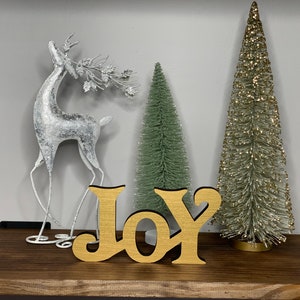 May include: A wooden sign with the word "JOY" in gold and black lettering, placed on a wooden shelf. The sign is accompanied by a silver reindeer figurine, two small Christmas trees, and a larger gold-colored tree, creating a festive holiday display.