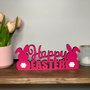 May include: Pink wooden sign with the text "Happy Easter" and two bunny silhouettes on either side. The sign has white accents on the bunnies.