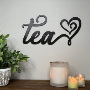 May include: Black wooden wall decor featuring the word "tea" in a cursive font and a stylised heart design. The decor is displayed on a white wall with a potted plant and candles on a wooden surface.