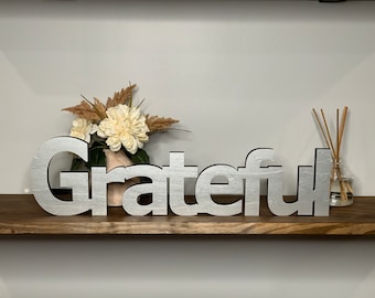 May include: Silver-painted wooden sign spelling out the word "grateful" on a wooden shelf. The sign is decorated with a vase of dried flowers.
