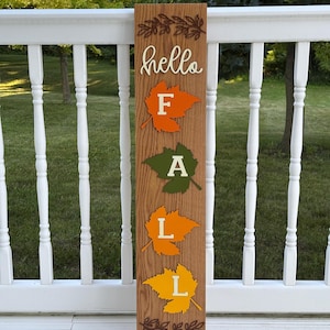 May include: A tall rectangular wooden sign with a natural wood grain finish. The sign features the word hello in white script at the top, followed by four stacked leaves in orange, green, yellow-orange, and yellow, each displaying a white letter F, A, L, L.