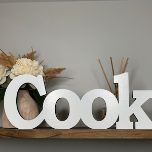 May include: White wooden letters spelling out the word "Cook" on a wooden shelf. The letters are large and have a slightly textured surface.