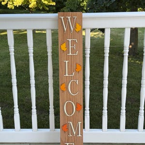 May include: A tall, rectangular wooden sign featuring the word WELCOME written vertically in white capital letters. Each letter is accented with a small, colorful cutout of an autumn leaf in shades of yellow, orange, and brown attached to the board.