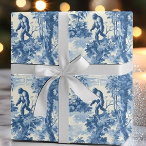 May include: A wrapped gift box featuring blue and white toile-style paper with a repeating pattern of a Bigfoot figure in a forest scene. The box is tied with a white satin ribbon and bow. Two lit candles are in the background.