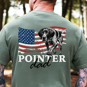 May include: A sage green t-shirt featuring a graphic of an American flag with a German Shorthaired Pointer dog. The text "POINTER dad" is printed below the flag and dog illustration.