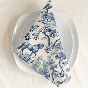 May include: A blue and white cotton napkin with a repeating pattern of horses and trees. The napkin is folded into a triangle and placed on a white plate.