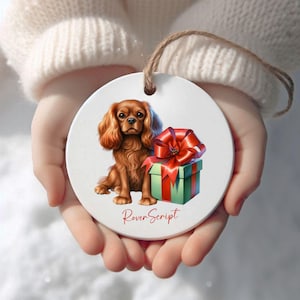 May include: A circular ceramic ornament with a Cavalier King Charles Spaniel illustration and a gift box with a red bow. The ornament has a brown string for hanging and the text "RoverScript" in red.