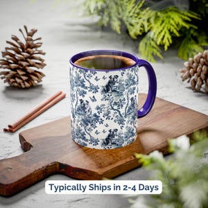 May include: A white ceramic mug with a blue and white floral pattern featuring a pug dog design. The mug has a purple rim and handle. The mug is sitting on a wooden chopping board.