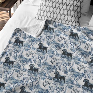 May include: A blue and white patterned throw blanket featuring a repeating design of black dogs, trees, and birds. The blanket has a soft, white sherpa lining on the underside. The blanket is displayed on a white bed with a patterned pillow.