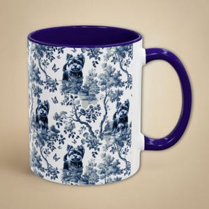 May include: A white ceramic coffee mug with a blue and white pattern of Yorkshire Terriers and floral designs. The mug has a purple rim and handle.