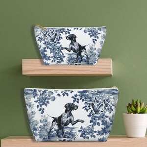 May include: Two toile-patterned zippered pouches with a white background and blue floral designs. The larger pouch features a black and white hunting dog with a blue collar. The smaller pouch features a similar design with a smaller dog.