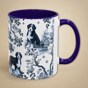 May include: A white ceramic mug with a deep blue interior and handle. The exterior features a repeating toile-style pattern in dark blue, depicting Bernese Mountain dogs, leafy trees, floral elements, and small butterflies on a white background.