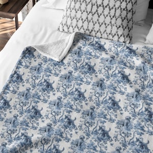 May include: A blue and white fleece throw blanket with a toile pattern featuring dogs and trees. The blanket has a white, fluffy underside and is draped over a bed with a patterned pillow.