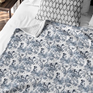 May include: A blue and white patterned throw blanket featuring a forest and dog design. The blanket is folded over a bed with a patterned pillow. The blanket has a soft, plush texture.