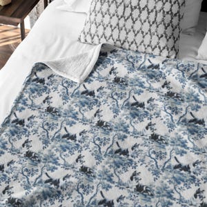 Op de afbeelding: Een witte deken met een blauw en wit toile de jouy patroon met bomen en dieren. De deken heeft een zachte, witte sherpa voering. De deken is opgevouwen over een bed met een kussen met patroon.
