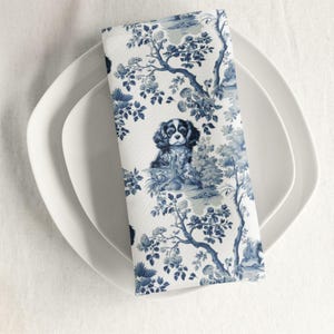 May include: Blue and white patterned fabric napkin with a blue and white illustration of a Cavalier King Charles Spaniel dog.