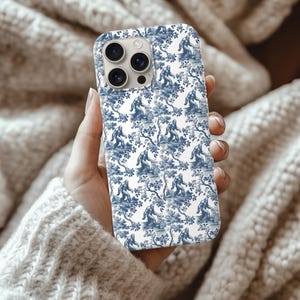 May include: A white phone case with a blue toile pattern featuring a Bigfoot design. The case has a glossy finish and a camera cutout. The design includes trees and Bigfoot figures in a repeating pattern.