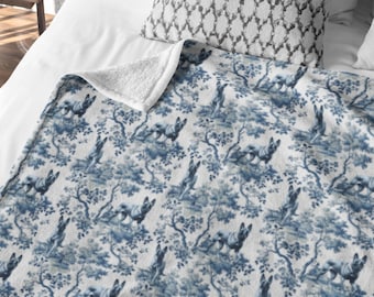 May include: A blue and white patterned throw blanket with a toile design of dogs and trees. The blanket has a soft, white sherpa lining on the underside. The blanket is folded over a bed with a patterned pillow.