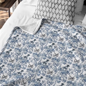 Pode incluir: Um cobertor azul e branco com um padrão toile de Jouy com cães e árvores. O cobertor tem um forro de sherpa branco e macio no verso. O cobertor está dobrado sobre uma cama com uma almofada estampada.