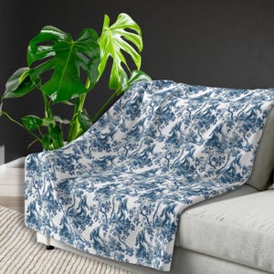 May include: A white throw blanket with a blue and white toile pattern featuring a repeating design of monkeys in trees. The blanket is draped over a white sofa with a green plant in the background.