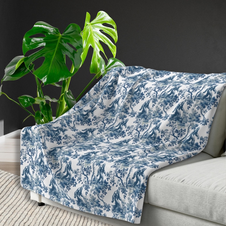 May include: A white throw blanket with a blue and white toile pattern featuring a repeating design of monkeys in trees. The blanket is draped over a white sofa with a green plant in the background.