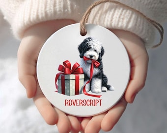 May include: A round, white ceramic ornament featuring a black and white dog with a red ribbon in its mouth, sitting next to a wrapped gift with a red bow. The ornament has a brown string for hanging and the word "ROVERSCRIPT" in red.