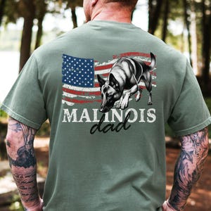 May include: Sage green t-shirt with a graphic of a Malinois dog and a distressed American flag. The text "MALINOIS dad" is printed below the flag and dog illustration.