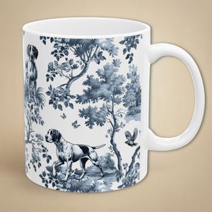 May include: White ceramic mug featuring a blue and white toile design with dogs, trees, and butterflies. The mug has a curved handle and a smooth, glossy finish. The design wraps around the entire mug.