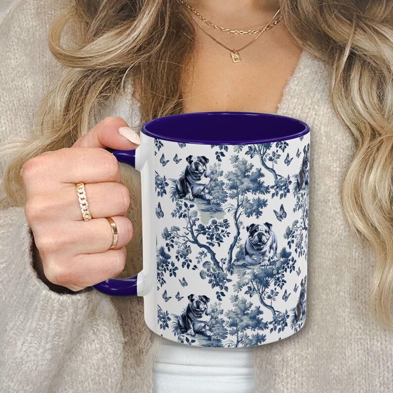 May include: White ceramic mug with a blue and white pattern of bulldogs and floral designs. The mug has a purple rim and handle.