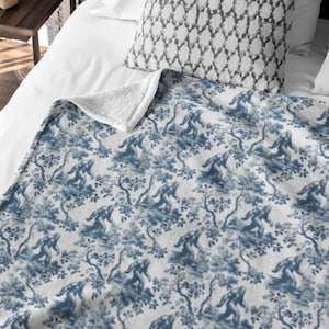 May include: A white and blue fleece throw blanket with a repeating pattern of blue Bigfoot figures walking through a forest. The blanket has a soft, plush texture.