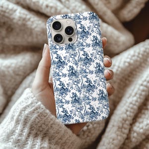 May include: A white phone case with a blue toile pattern featuring figures fishing near trees. The case has a glossy finish and a camera cutout. The case is held in a hand.