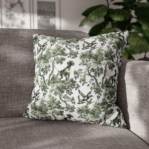 May include: Square cushion with a green and white toile design depicting dogs, trees, and flying ducks. The cushion is placed on a grey sofa, adding a decorative touch to the living space.