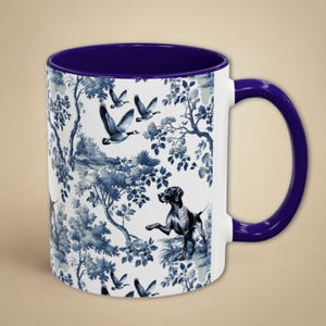 May include: A white ceramic mug with a blue and white toile pattern featuring a dog and birds. The mug has a blue rim and handle.