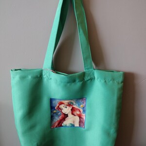 Peut inclure: Un sac fourre-tout vert avec de longues anses. Le sac pr&eacute;sente une image color&eacute;e d'un personnage de dessin anim&eacute; sur le devant. Le personnage a de longs cheveux roux et porte un haut bleu. Le sac est suspendu contre un mur gris.