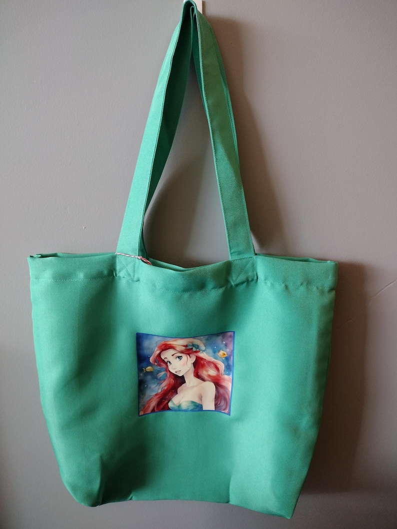 Peut inclure: Un sac fourre-tout vert avec de longues anses. Le sac pr&eacute;sente une image color&eacute;e d'un personnage de dessin anim&eacute; sur le devant. Le personnage a de longs cheveux roux et porte un haut bleu. Le sac est suspendu contre un mur gris.