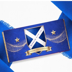 May include: A blue chocolate bar with gold trim and accents. The bar features a shield with a white cross and the text "WORLD CUP '26". Gold stars and the text "MANDATORY MATCH CHOCOLATE" are also visible on the bar.