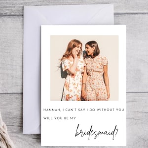 May include: A white card with a photo of two women smiling at each other. The text on the card reads "HANNAH, I CAN'T SAY I DO WITHOUT YOU WILL YOU BE MY bridesmaid?"