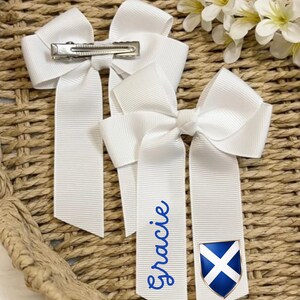 May include: Three white hair bows with long ribbons. One ribbon has the name "Gracie" in blue script. Another ribbon features a blue and white shield emblem. Each bow has a metal clip on the back. The bows are on a woven background.