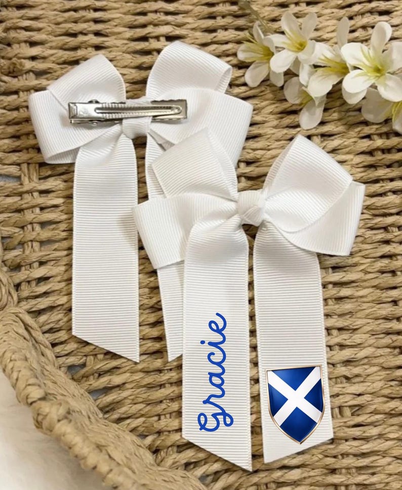 May include: Three white hair bows with long ribbons. One ribbon has the name "Gracie" in blue script. Another ribbon features a blue and white shield emblem. Each bow has a metal clip on the back. The bows are on a woven background.