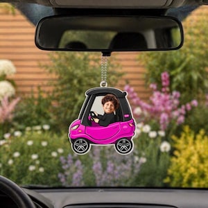 May include: A pink car-shaped air freshener with a smiling child inside, hanging from a car's rearview mirror. The car has black wheels and a black roof. The background is a blurred outdoor scene with flowers and greenery.