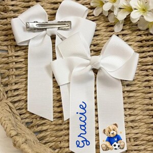 May include: Three white hair bows with a grosgrain ribbon design. One bow has a silver clip, and two have long tails. One tail is personalised with the name "Gracie" in blue script, and the other has a teddy bear graphic wearing a blue football jersey.