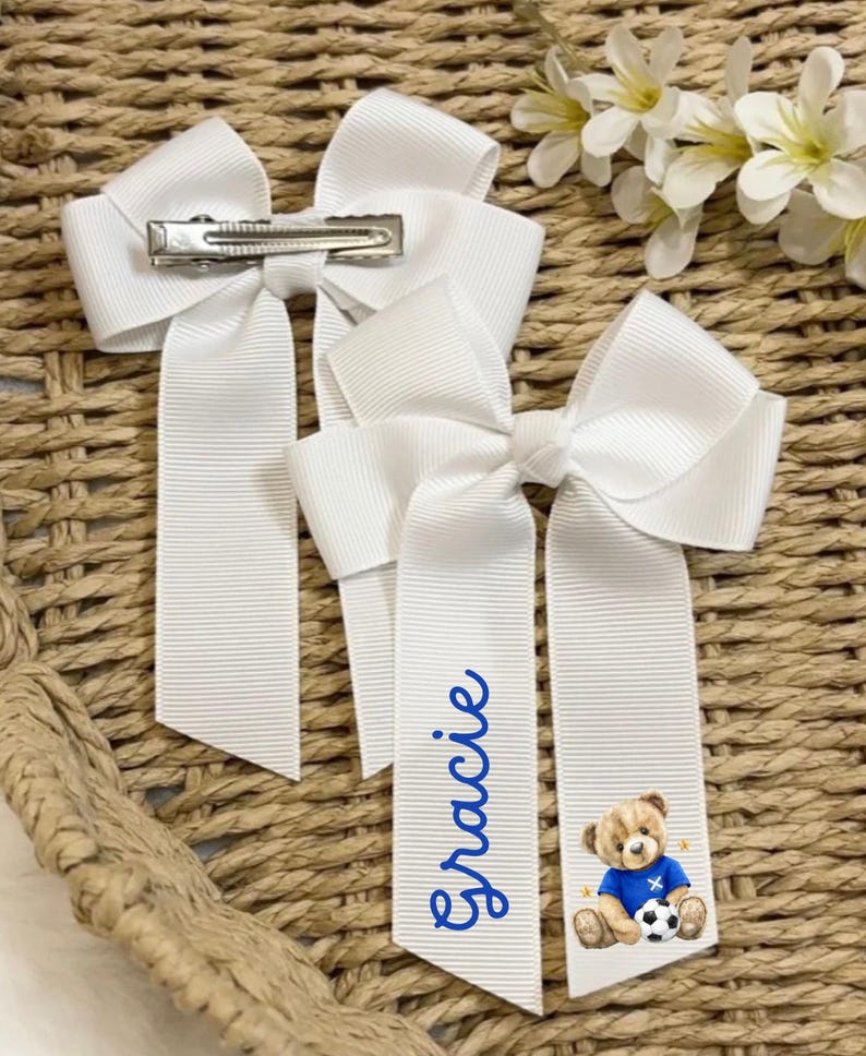 May include: Three white hair bows with a grosgrain ribbon design. One bow has a silver clip, and two have long tails. One tail is personalised with the name "Gracie" in blue script, and the other has a teddy bear graphic wearing a blue football jersey.