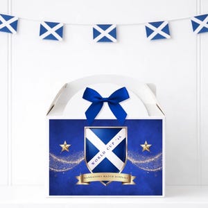 May include: A white box with a blue bow, adorned with a Scottish flag motif. The box displays a shield with the Scottish flag, gold stars, and the text "WORLD CUP '16" and "MANDATORY MATCH SUPPLIES". A garland of Scottish flags is suspended above.