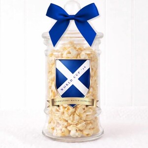 May include: A glass jar filled with popcorn, adorned with a blue bow. The jar features a label with a Scottish flag design, the text "WORLD CUP '16", and "MANDATORY MATCH SUPPLIES".