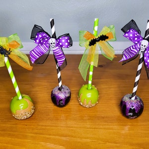 May include: Four Halloween-themed caramel apples with different colored bows and straws. The apples are decorated with glitter and caramel. The bows are black, purple, and green.