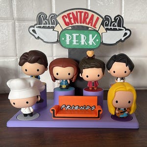 May include: A Central Perk display featuring six stylized figures of characters from the TV show Friends. The display includes a purple base, an orange couch with the word "FRIENDS", and a Central Perk sign.