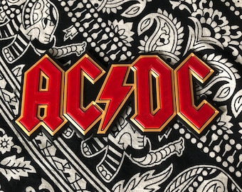 May include: A red and black metal sign with the word "AC/DC" in bold letters. The letters are outlined in gold.